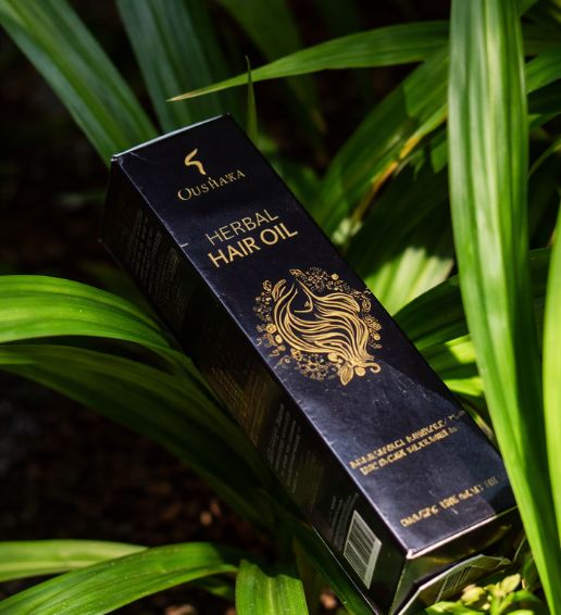 A 190ml bottle of Oushara Herbal Hair Oil with a minimalist cream label, elegantly placed on a piece of natural driftwood against a soft green background with a palm leaf.
