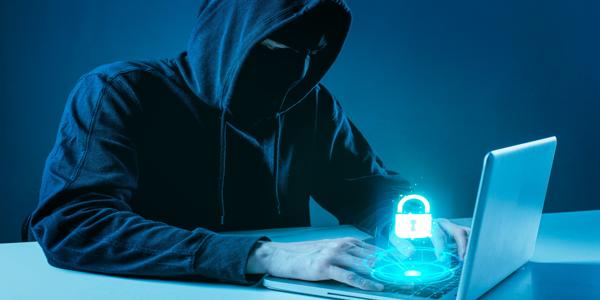 A person wearing a dark hoodie and mask typing on a laptop, symbolizing cybersecurity and data protection with a glowing digital padlock icon on the keyboard