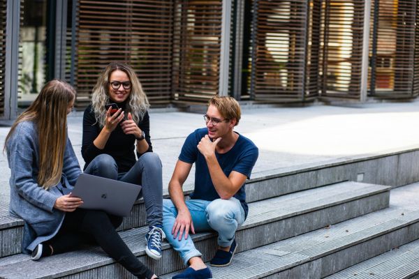 Three young adults sitting on outdoor steps, talking and using a laptop and smartphone.