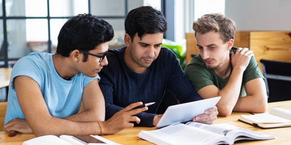 Three young men sitting at a table studying together while looking at a tablet.