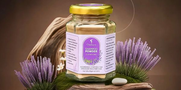 A glass jar of Oushara Lavender Shampoo Powder placed on a zen-like arrangement of mossy stones and driftwood, surrounded by sprigs of fresh lavender and small white flowers.