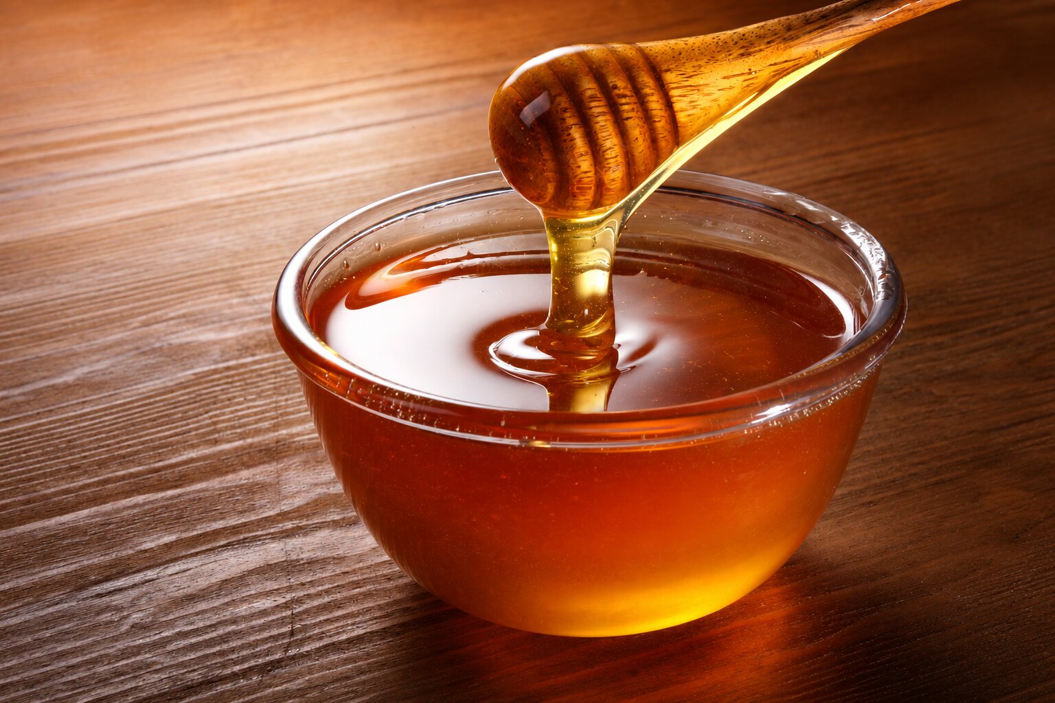 Thick golden honey dripping from a wooden dipper into a clear glass bowl, set on a warm wooden tabletop with studio lighting.